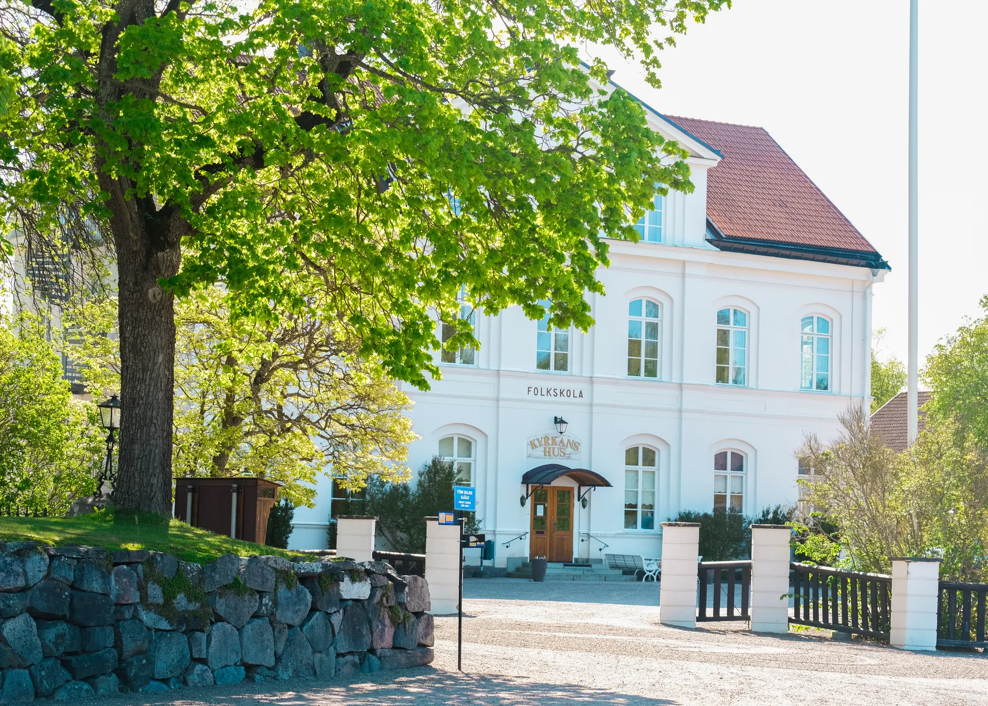 En vit tvåvåningsbyggnad med texten FOLKSKOLA på fasaden, framför ett stort lövträd och en stenmur en solig vårdag.