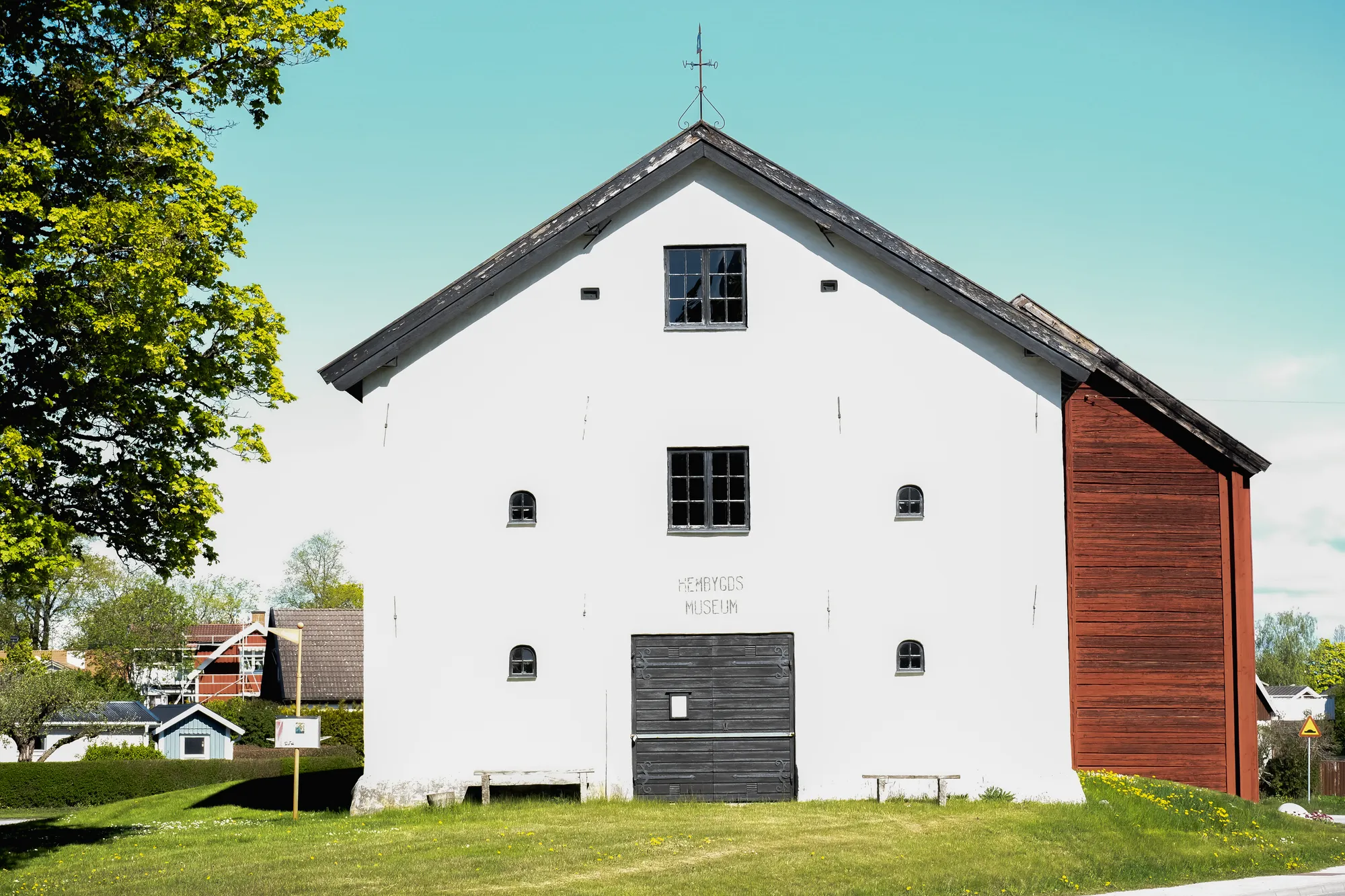 En stor vitkalkad magasinsbyggnad med rött trätillbygge och texten HEMBYGDS MUSEUM på gaveln, omgiven av grön gräsmatta.