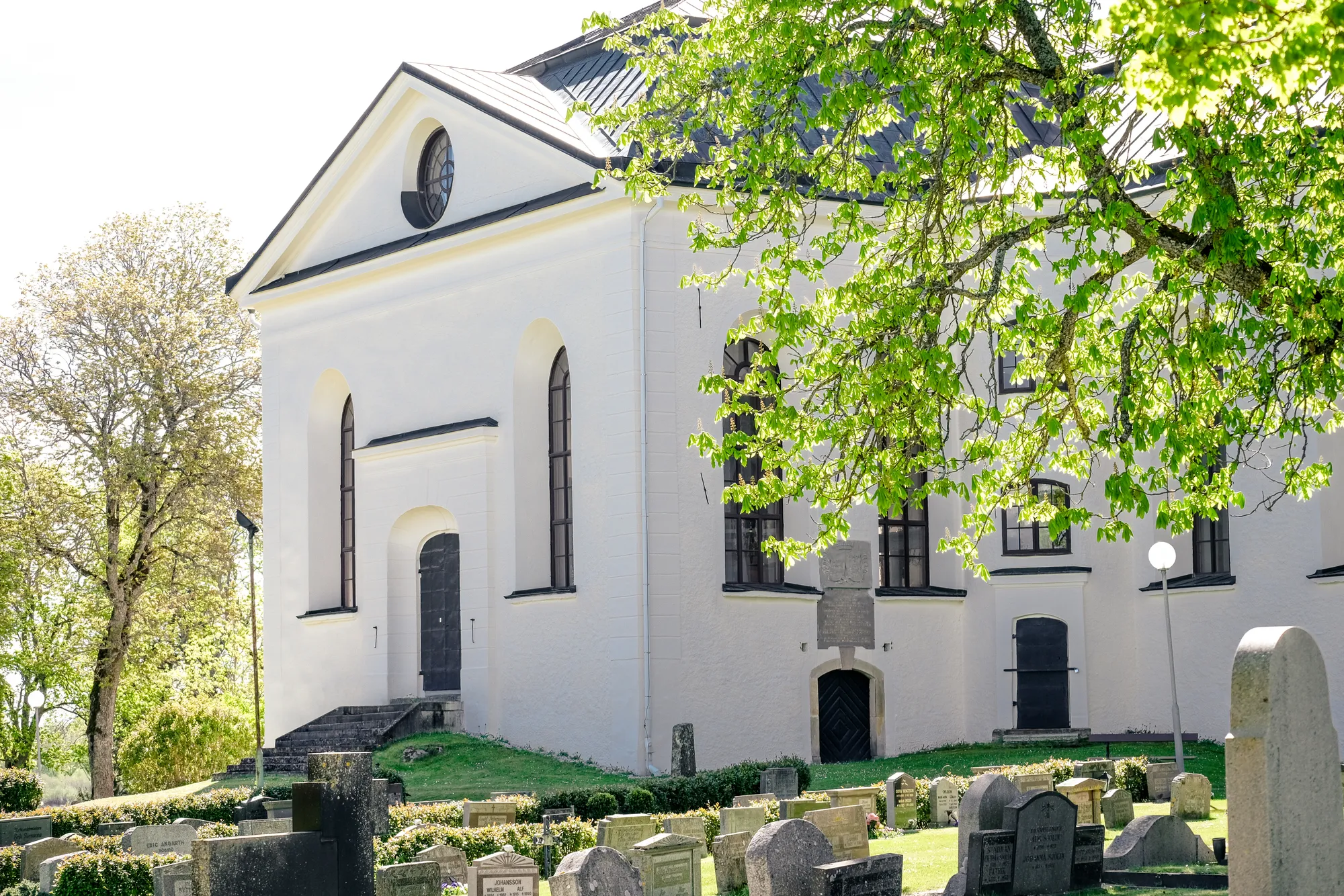 En vitputsad kyrka med rund fönsterros och spetsbågefönster, omgiven av gravstenar på en kyrkogård och grönskande lövträd.
