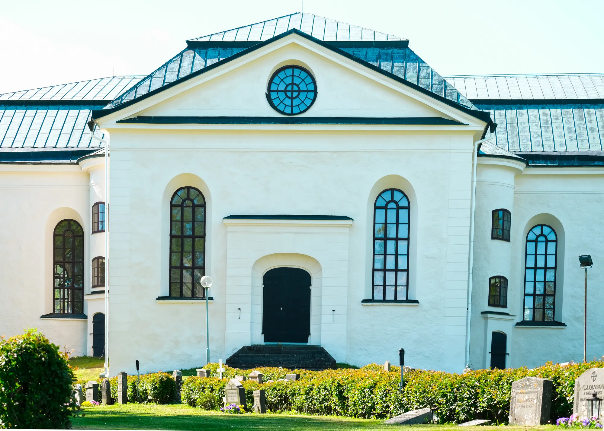 Gaveln på en vitputsad kyrka med rund fönsterros, rundbågefönster och svart port, med gravstenar och häckar framför.