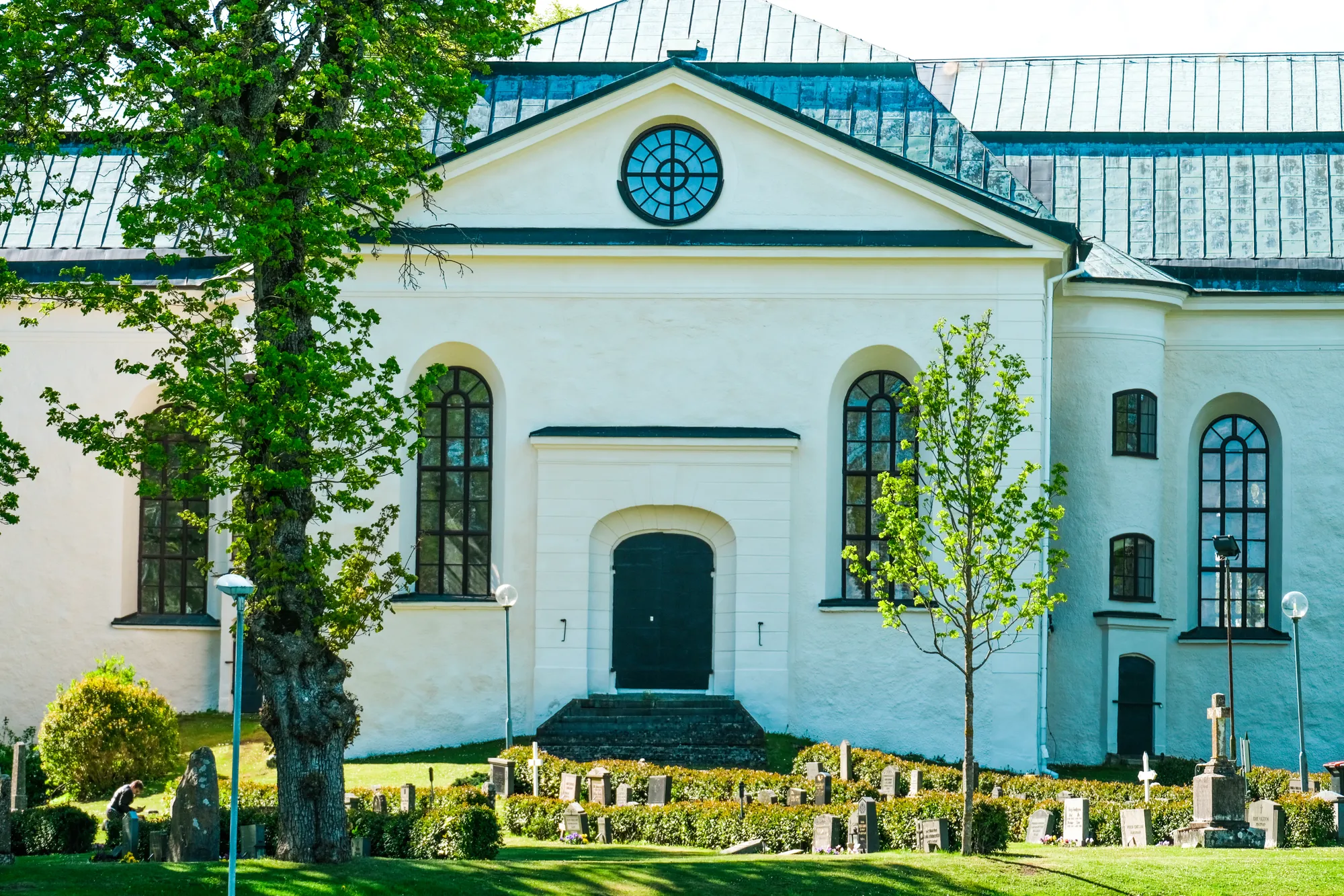 Korsarmen på en vit kyrka med rund fönsterros och svart port, med gravstenar, häckar och ett stort lövträd framför.