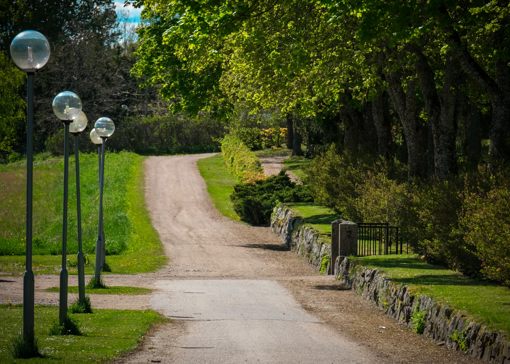 En grusväg kantad av runda parklyktor och en låg stenmur slingrar sig genom grönskande lövträd en solig dag.