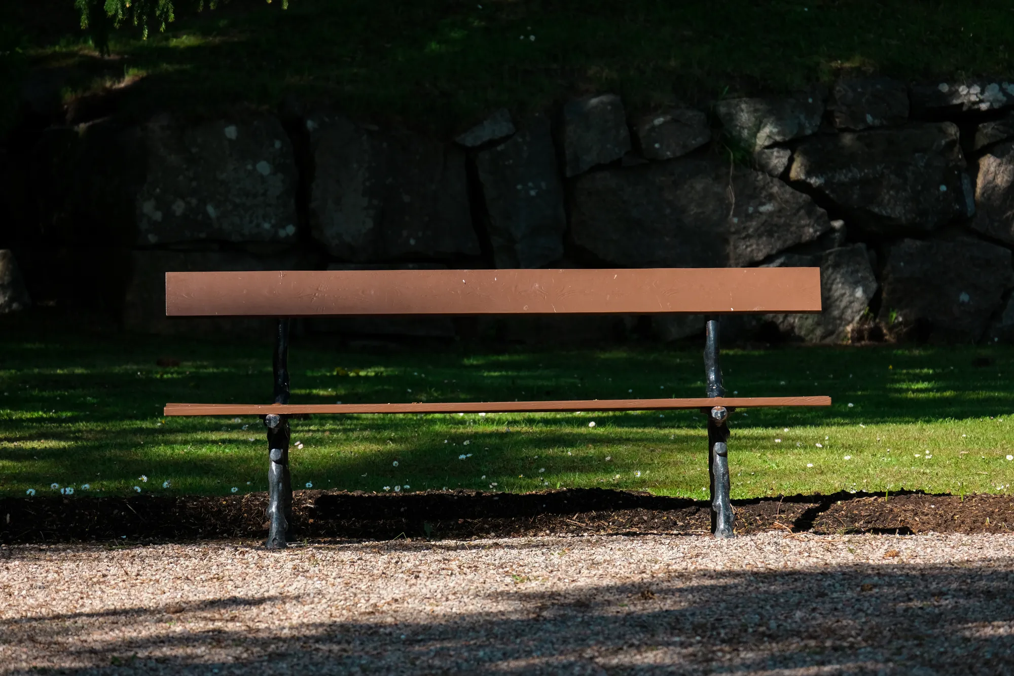 En tom parkbänk i trä står på en grusgång framför en stor stenmur, delvis solbelyst.