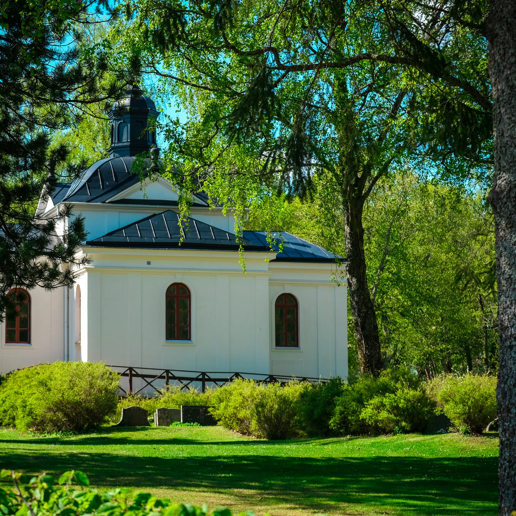 Ett vitt åttkantigt kapell med kupol och svart tak skymtar bakom grönskande lövträd och buskar i en park.