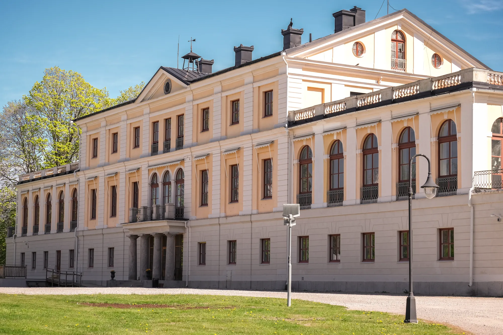 En stor gulvit herrgårdsbyggnad i två våningar med valvfönster, sedd snett från sidan med gräsmatta och en lyktstolpe framför.