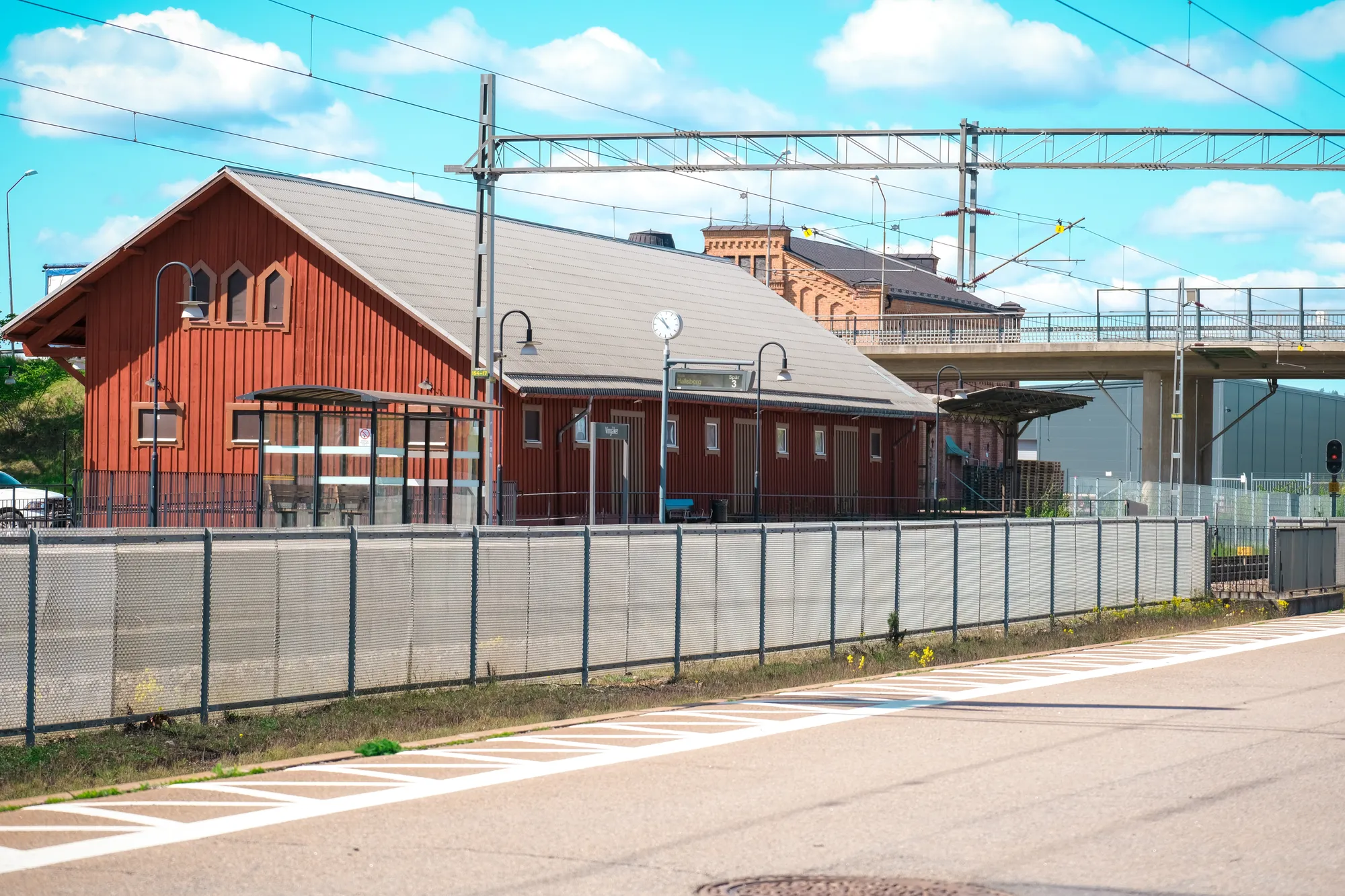 En röd stationsbyggnad i trä bredvid järnvägsspår med kontaktledningar och en betongviadukt i bakgrunden.