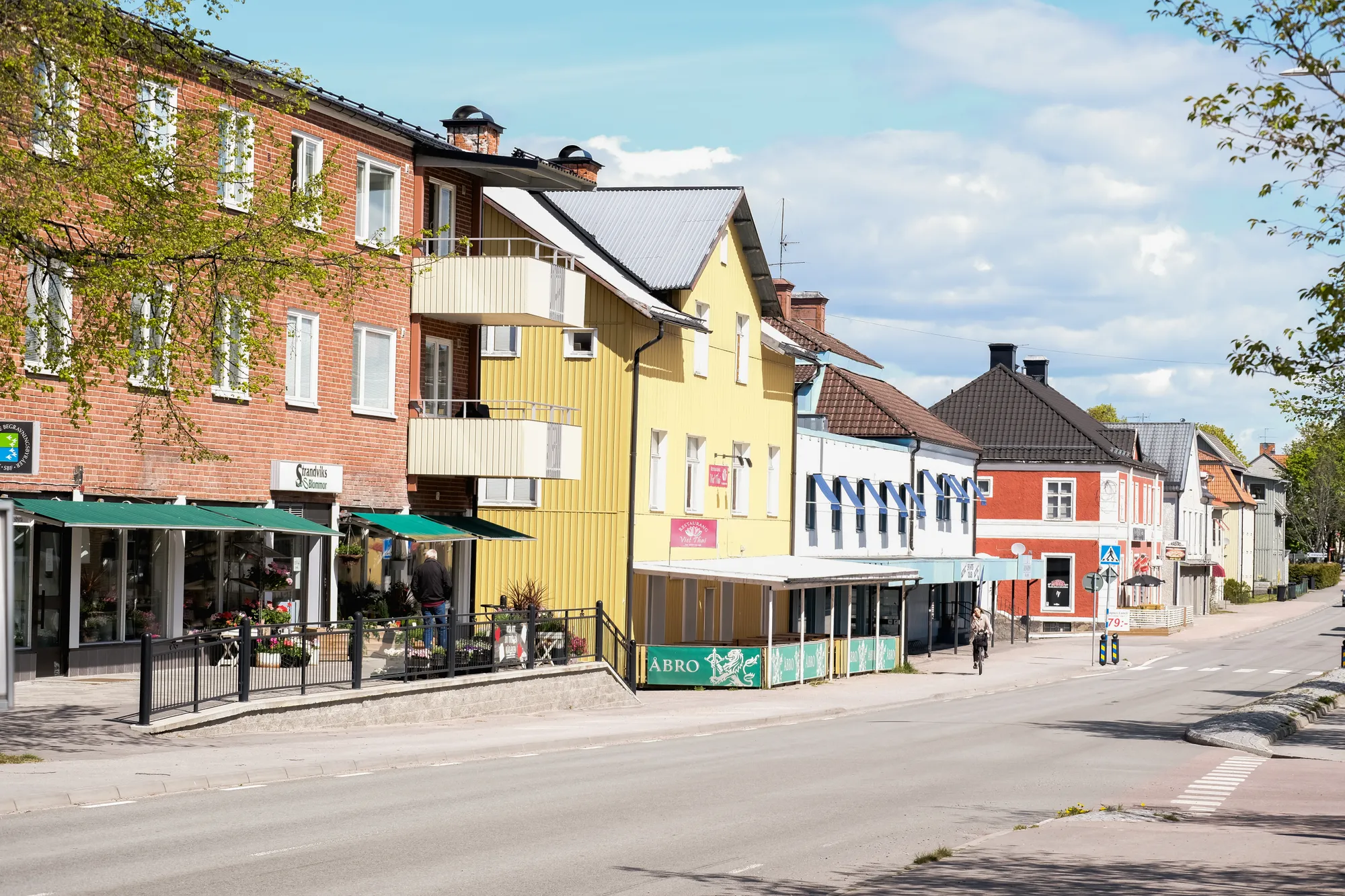 En lugn gata i tätort med flerbostadshus, butiker och en gul träbyggnad längs vägen.