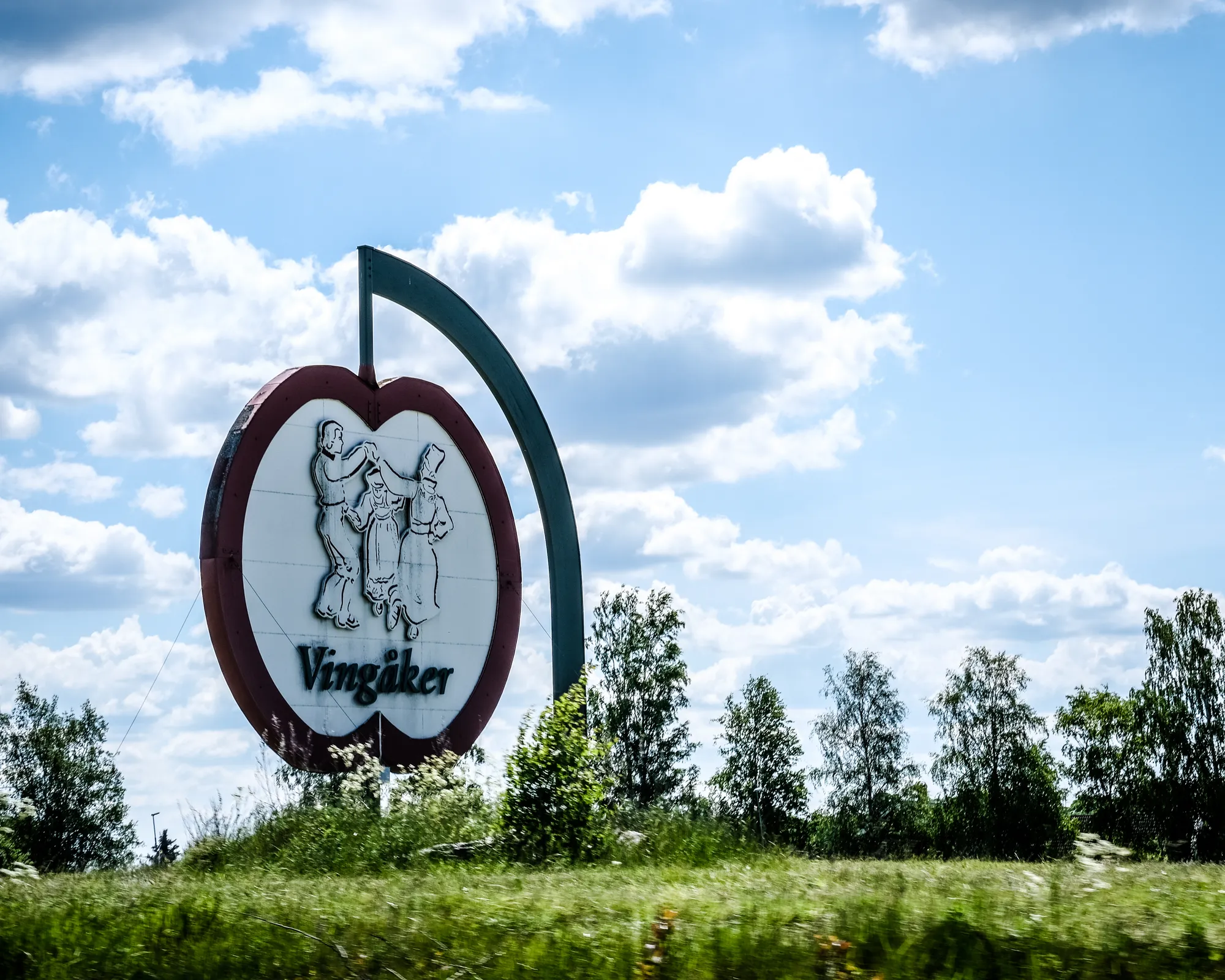 En stor skylt formad som ett äpple med dansande folkdräktsklädda figurer och texten Vingåker, placerad på en gräsbevuxen kulle mot molnig himmel.