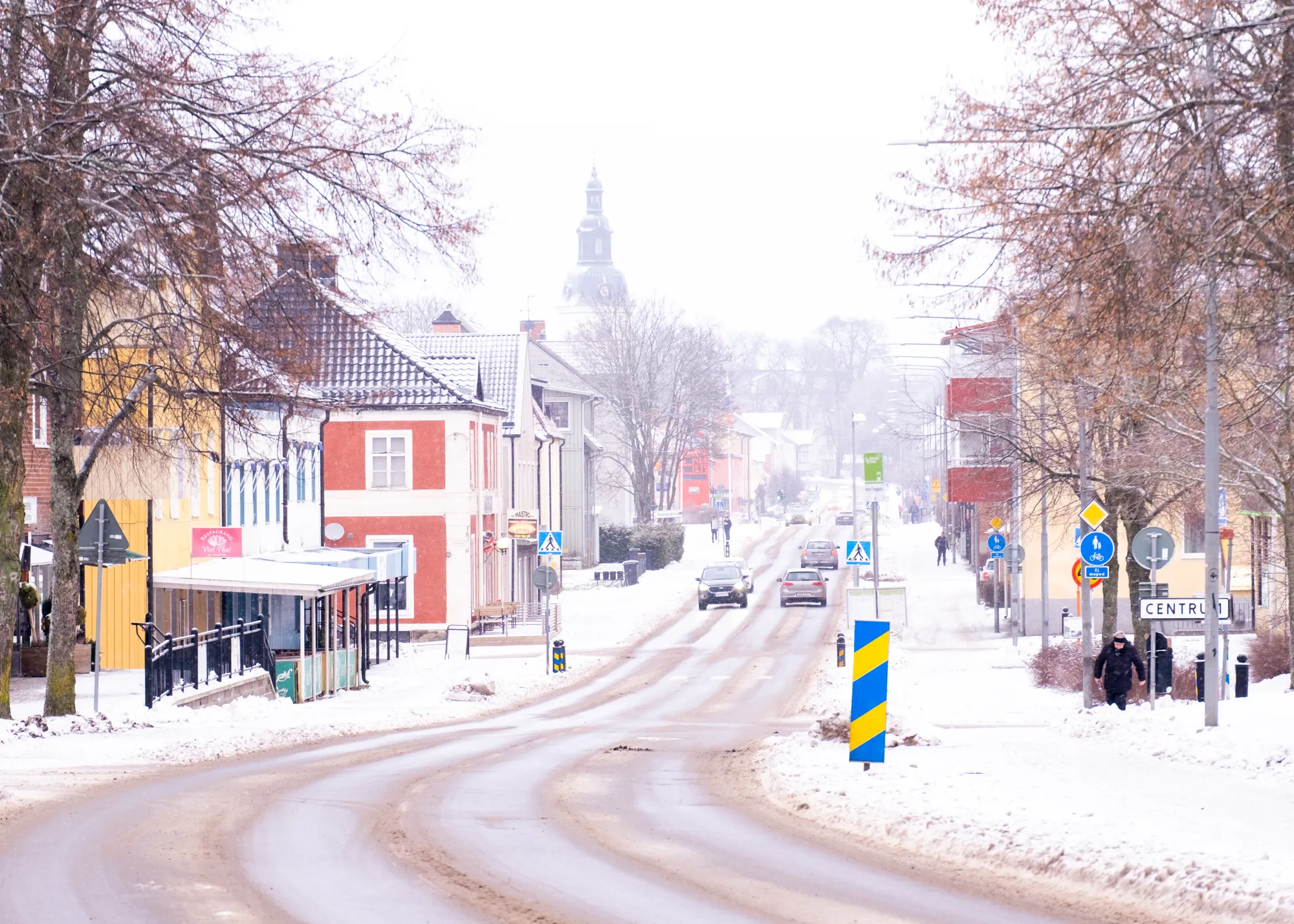 En snöig huvudgata genom ett samhälle med bilar, en kyrktorn i fjärran och en Centrum-skylt.