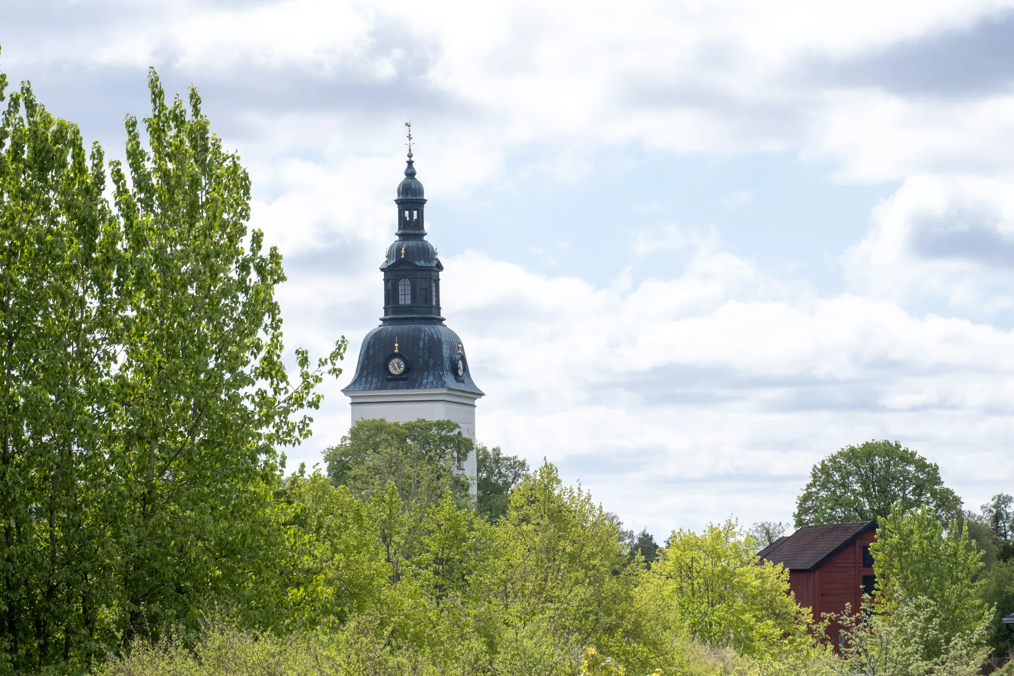 Ett svart kyrktorn med spira reser sig bakom grönskande lövträd under en himmel med moln, med en röd byggnad till höger.