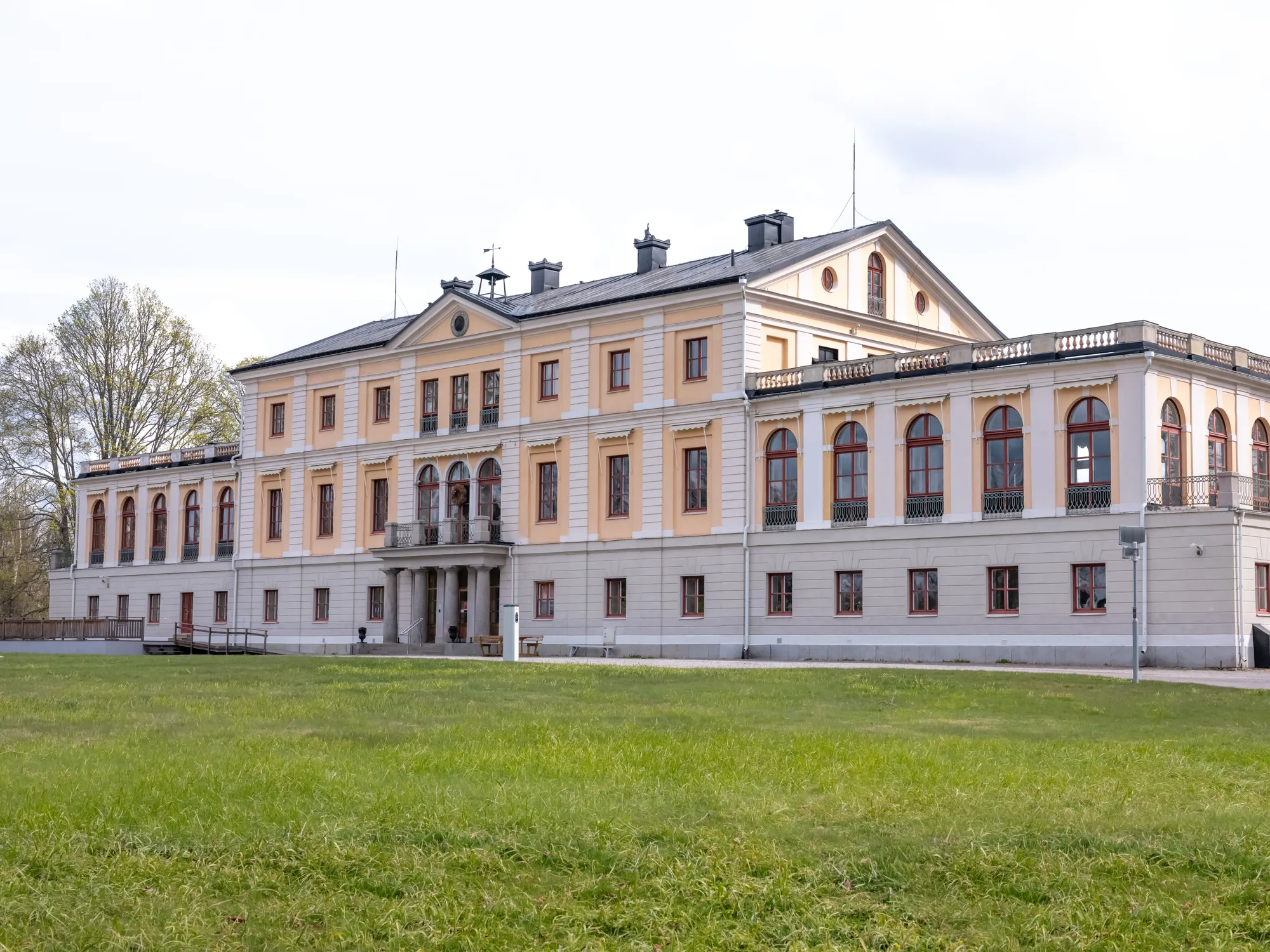 En stor gulvit herrgård med flera fönsterrader och svart tak sedd snett framifrån med en gräsmatta i förgrunden.