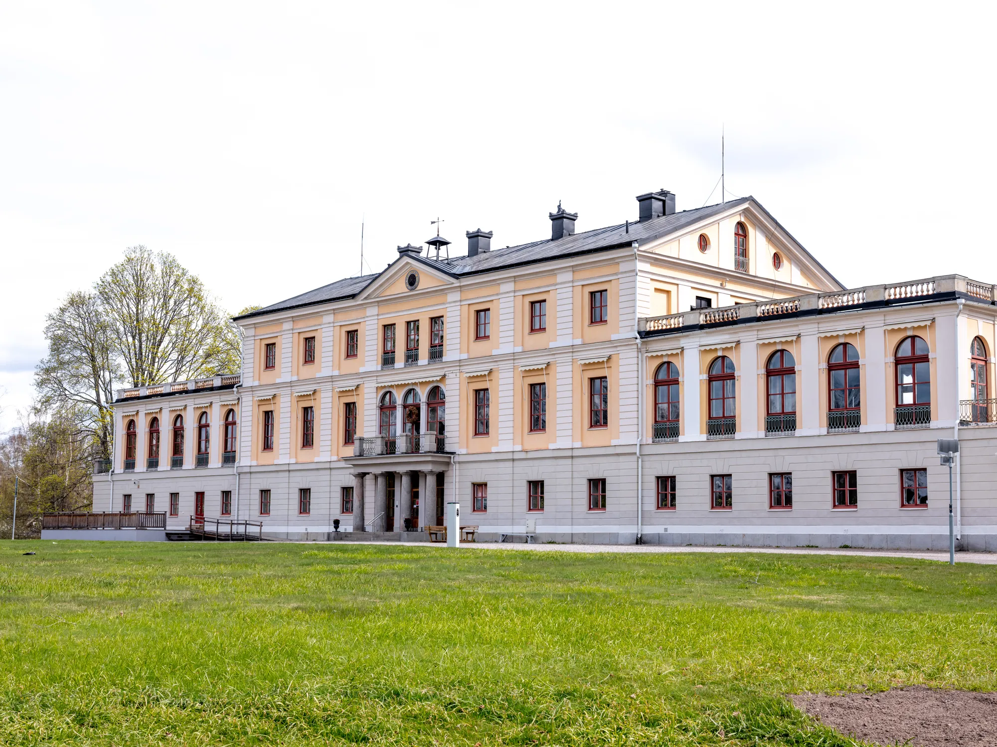En gulvit herrgård med valvfönster och balkong sedd snett framifrån med en grön gräsmatta i förgrunden.