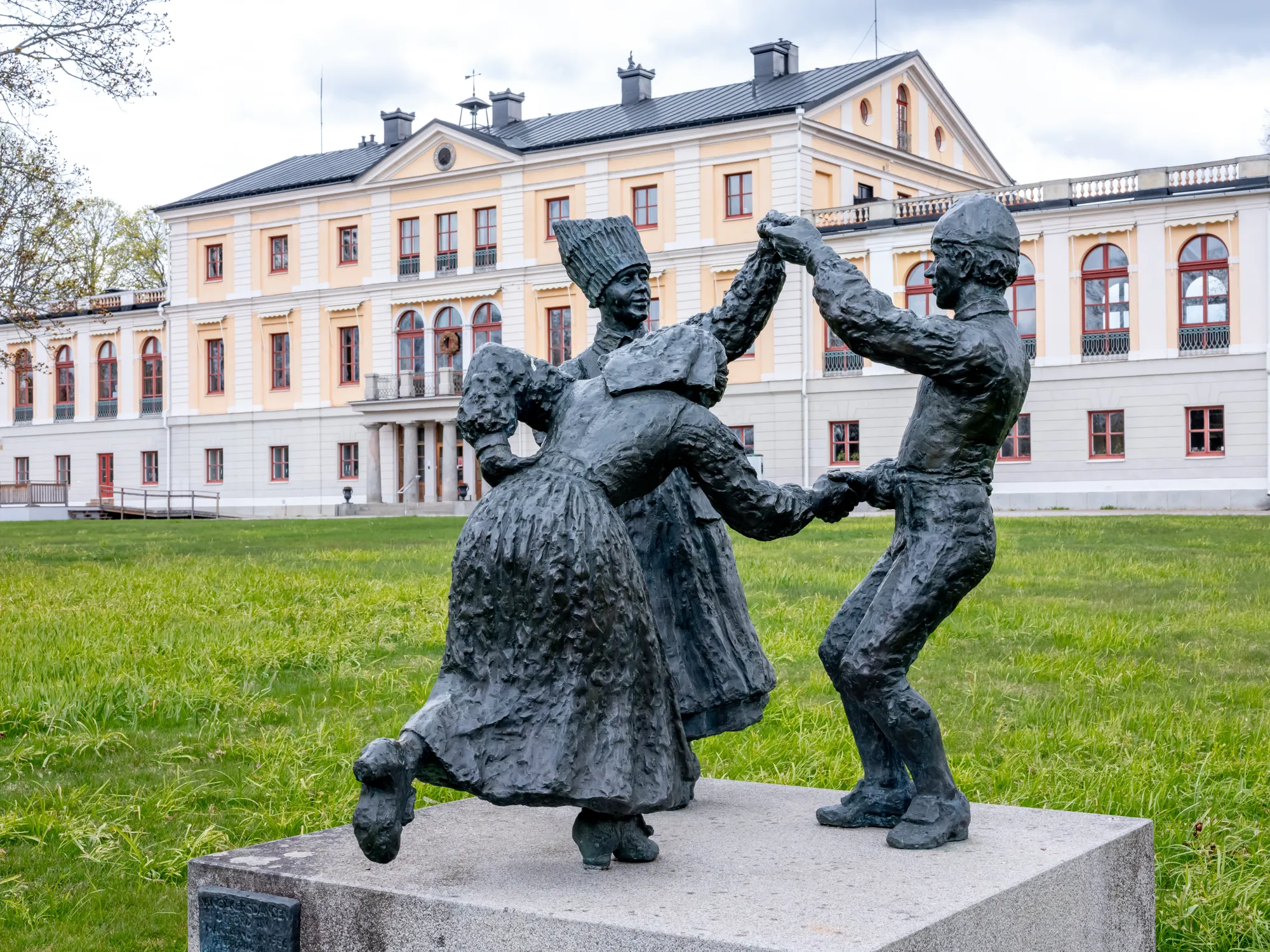 En bronsstaty av ett dansande par i folkdräkt på en stensockel framför en gulvit herrgård.