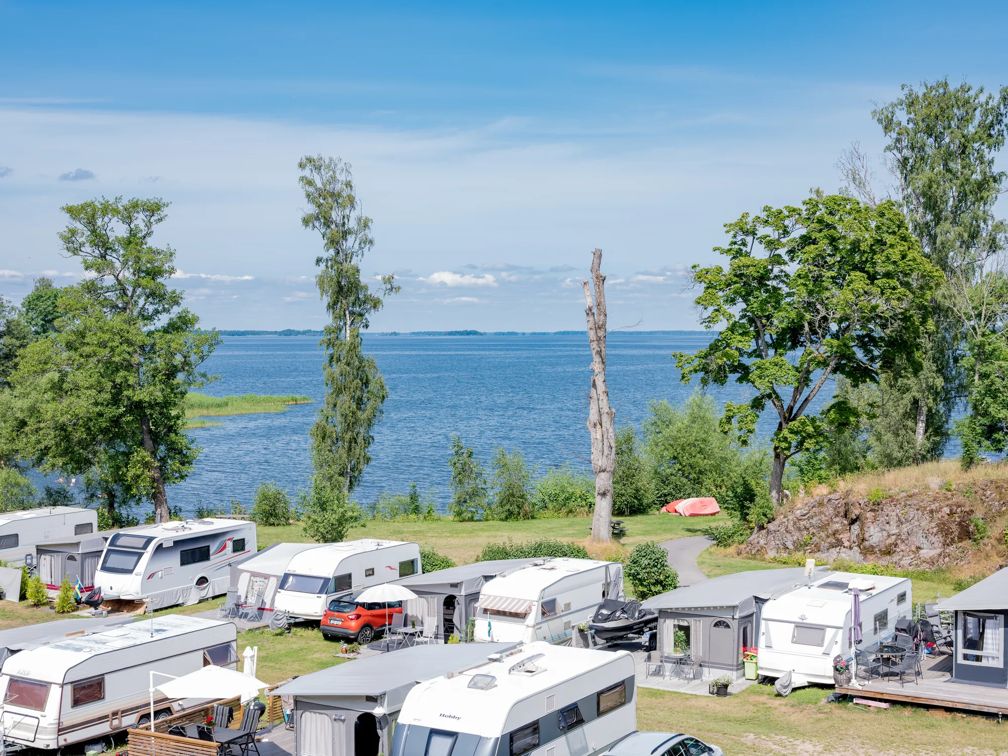 En campingplats med många husvagnar och förtält på gräsmark vid en stor sjö under en blå himmel.