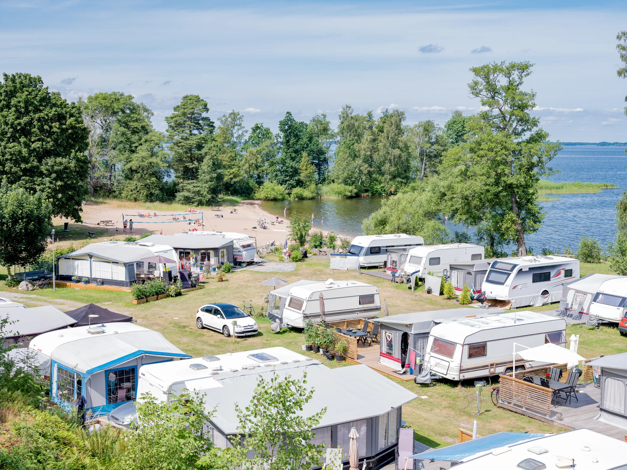 En översiktsbild över en campingplats med husvagnar och förtält intill en badstrand och en stor sjö under en blå himmel.